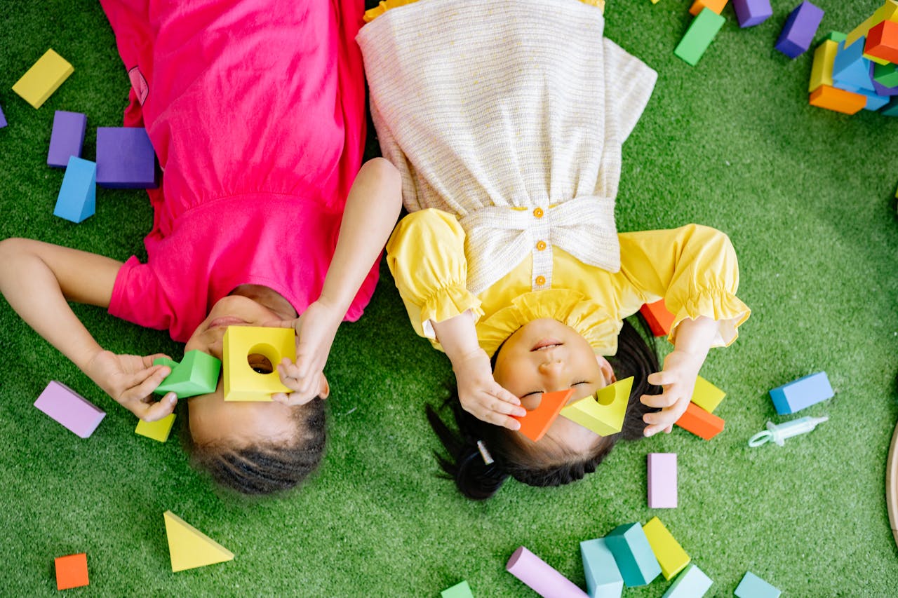 Two joyful kids playing with colorful wooden blocks on a green surface indoors.